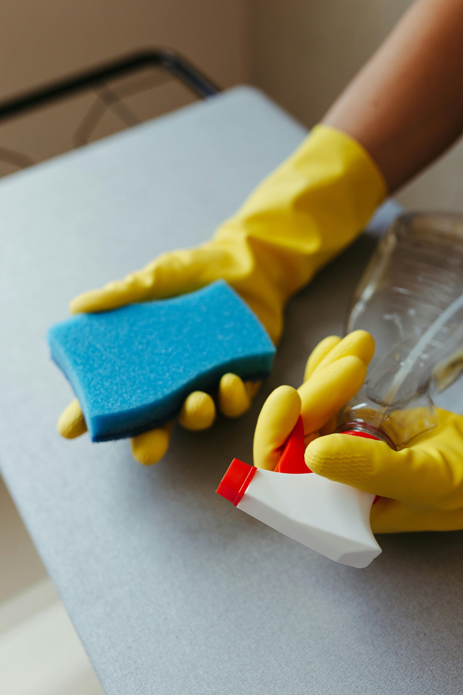 Gros plan sur une personne nettoyant une surface avec une éponge bleue et un vaporisateur, portant des gants jaunes.