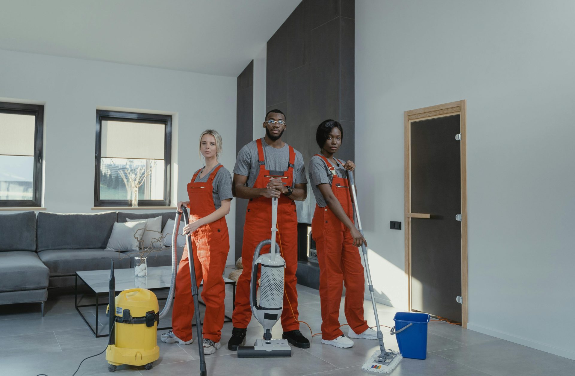 Une équipe de nettoyage diversifiée, vêtue d'uniformes rouges, prête à désinfecter un salon moderne à l'aide d'un aspirateur et d'une serpillière.