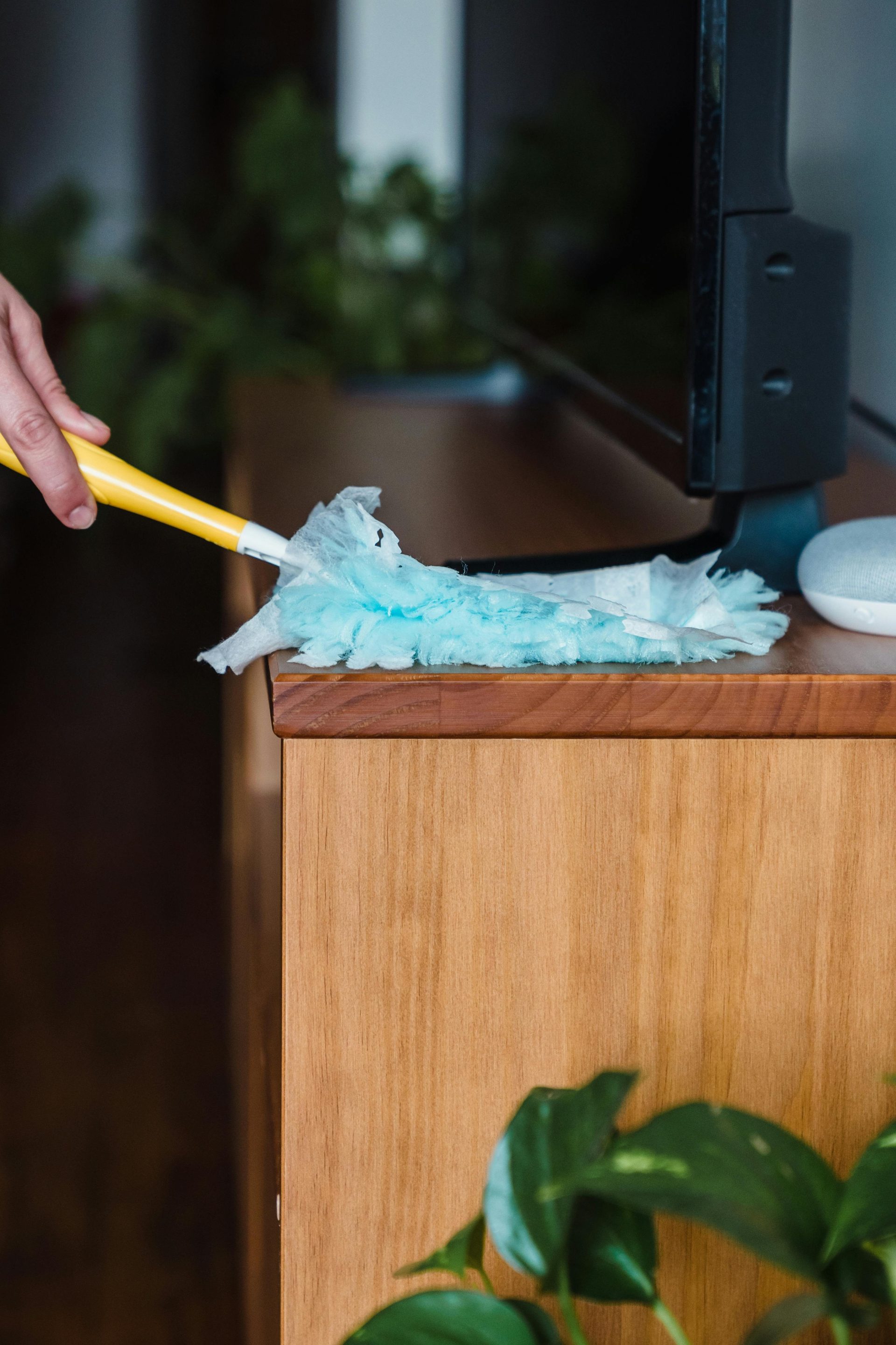 Close-up of a person using a blue duster on a wooden surface near a TV indoors.